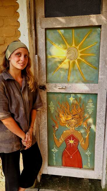 A smiling woman with a green headband stands beside a door featuring vibrant sun and goddess artwork, blending earthy, spiritual, and artistic vibes in natural light.