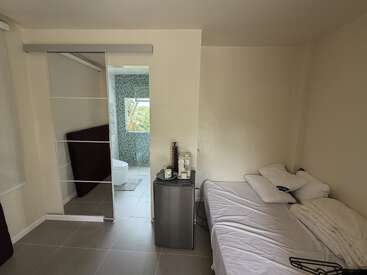 A small, minimalist room with a bed, compact fridge, coffee maker, glass sliding closet door, and tiled bathroom with a window. Clean and simple design.
