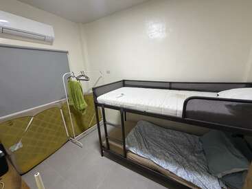 This image shows a small, tidy bedroom with a bunk bed, air conditioning, a window shade, clothing rack, and minimal furniture. The room looks functional.