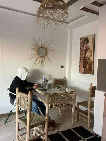A person in a hoodie works on a laptop at a rustic table with woven chairs. The room features earthy decor, a mirror, and a painting.