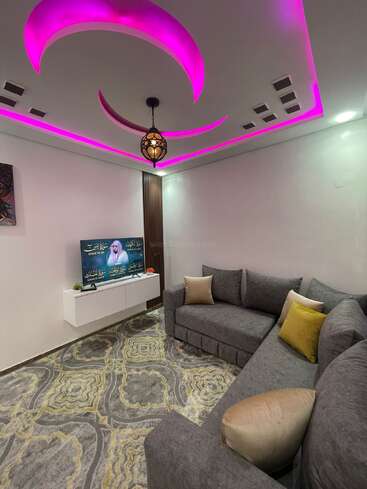 This modern living room features a grey sectional sofa with gold cushions, a patterned rug, floating TV stand, and dynamic pink LED ceiling lights with a hanging lamp.