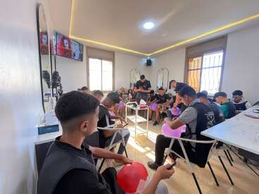 A group of young men sits in a brightly lit room, each holding balloons and pens, appearing focused on an activity, likely in a classroom or workshop setting.