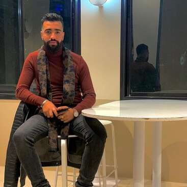 A bearded man sits confidently on a chair by a white table indoors, wearing a burgundy sweater, dark scarf, blue jeans, and holding a phone.