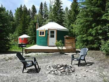 A cozy green yurt with a small porch sits among tall pine trees. A fire pit, grill, and three black chairs create a welcoming campsite.