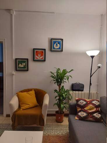 Uma sala de estar aconchegante com poltrona branca, travesseiro amarelo, almofada de sofá estampada, luminária de chão, planta de interior, piso de ladrilho e três obras de arte coloridas emolduradas na parede.