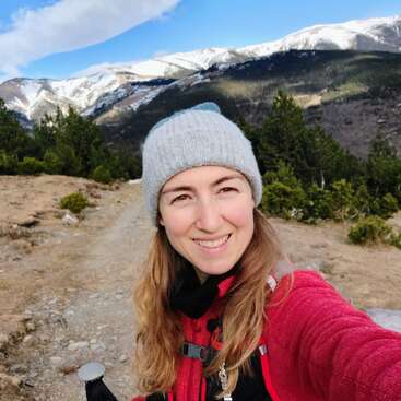 Uma mulher sorridente, usando um gorro cinza e uma jaqueta vermelha, tira uma selfie em uma trilha na montanha. Os picos cobertos de neve e as árvores verdes preenchem o belo plano de fundo.