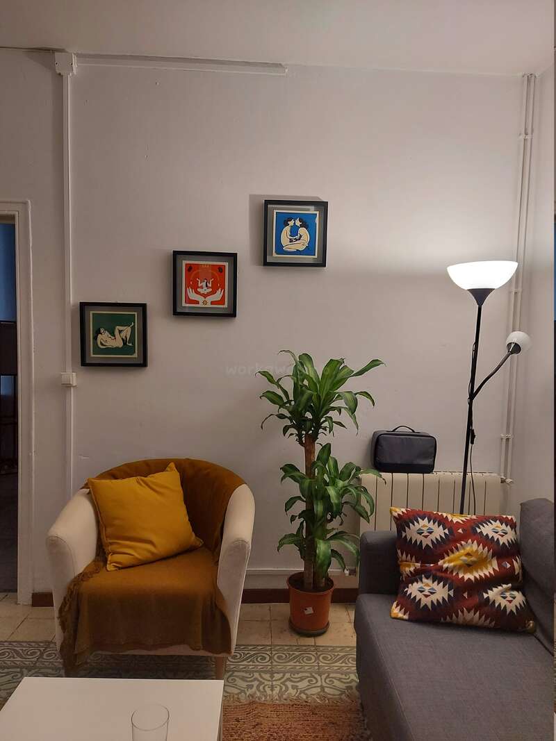 Uma sala de estar aconchegante com poltrona branca, travesseiro amarelo, almofada de sofá estampada, luminária de chão, planta de interior, piso de ladrilho e três obras de arte coloridas emolduradas na parede.