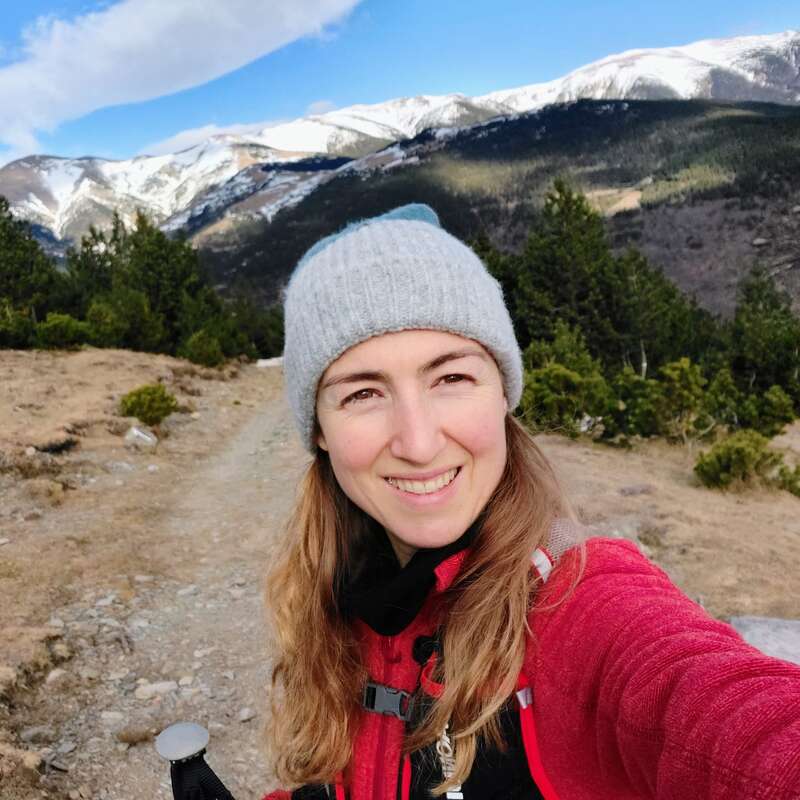 A smiling woman wearing a gray beanie and red jacket takes a selfie on a mountain trail. Snow-capped peaks and green trees fill the beautiful background.