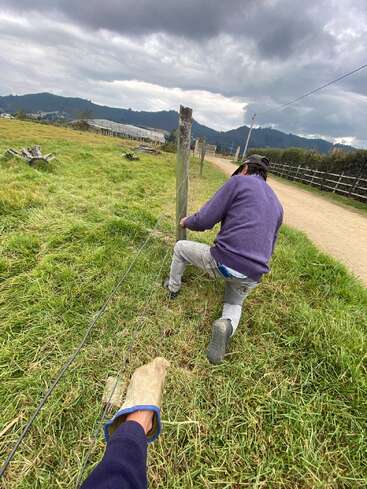 Une personne est agenouillée sur l'herbe, occupée à travailler sur un poteau de clôture métallique, tandis qu'une autre tient le fil de fer d'une main gantée. En arrière-plan, on aperçoit la campagne, les montagnes et un ciel nuageux.