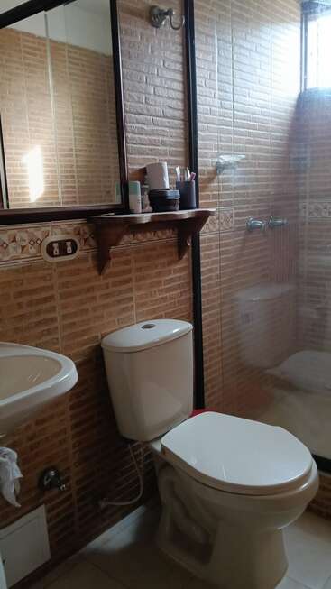 Cette photo montre une salle de bains avec des murs carrelés de couleur marron, des toilettes blanches, un lavabo, une cabine de douche en verre, un miroir, une étagère et divers articles de toilette.