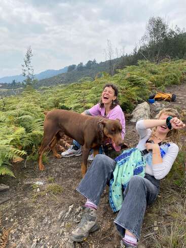 Deux femmes sont assises par terre à l'extérieur et rient joyeusement, un chien brun à leurs côtés. Elles sont entourées de fougères vertes et de montagnes lointaines, sous un ciel nuageux.