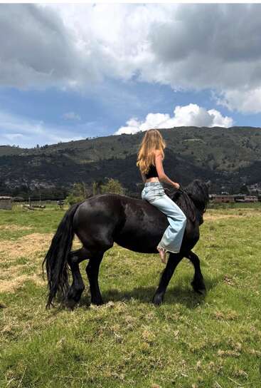 Une femme aux longs cheveux blonds monte à cheval, pieds nus, vêtue d'un haut noir et d'un jean bleu clair, au milieu d'un paysage de prairies verdoyantes et de montagnes.