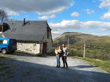 L'image représente une scène sereine d'un couple se tenant devant une maison rustique en pierre, sur un fond pittoresque de collines ondulantes et d'un ciel bleu avec des nuages blancs.