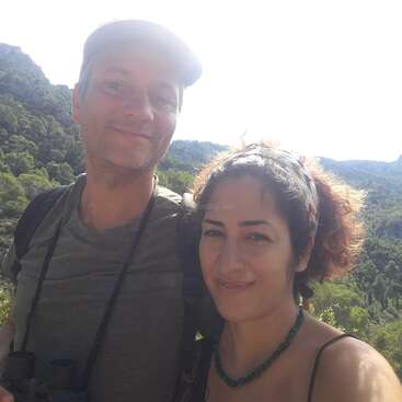 The image depicts a man and woman posing for a selfie in front of a mountainous landscape, with the woman wearing a headband and necklace.