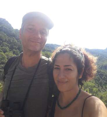 Das Bild zeigt einen Mann und eine Frau, die für ein Selfie vor einer Berglandschaft posieren, wobei die Frau ein Stirnband und eine Halskette trägt.