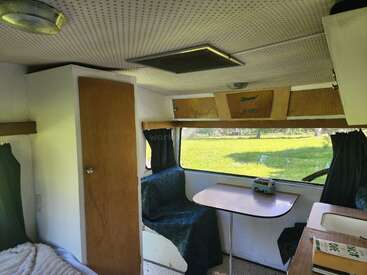This image shows the cozy interior of a camper van, featuring a small dining table, cushioned seating, green curtains, a wooden door, and overhead storage.