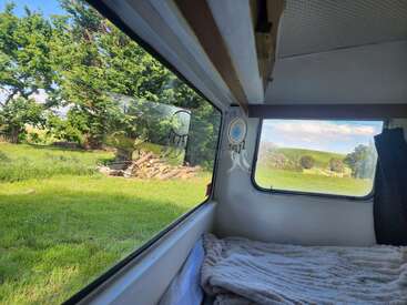 This image shows a cozy bed inside a camper van, looking out through large windows at lush green grass, trees, and a peaceful countryside landscape.