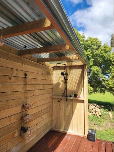 This image shows an outdoor wooden shower with a corrugated metal roof, hanging woven baskets, wooden shelves, and a beautiful green yard with trees and blue sky.