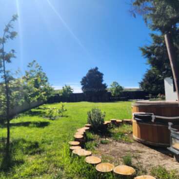 A sunny backyard with lush green grass, wooden fence, scattered trees, and a wooden hot tub bordered by circular stepping stones, under a bright clear blue sky.