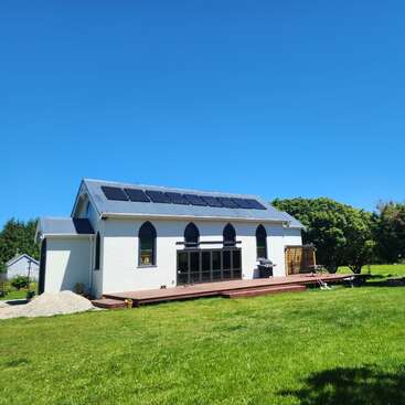 Um edifício branco moderno com grandes janelas em arco, painéis solares no telhado, deck de madeira, cercado por grama verde e árvores sob um céu azul claro.