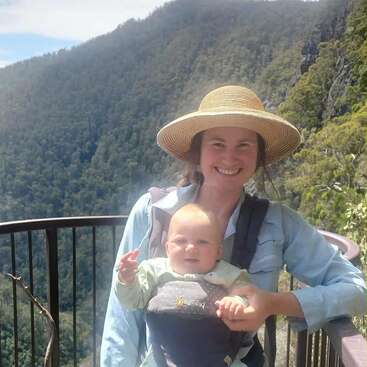 Une femme souriante portant un chapeau de soleil tient un bébé dans un porte-bébé. Elles se tiennent sur un point de vue panoramique avec des montagnes verdoyantes et un ciel bleu derrière elles.