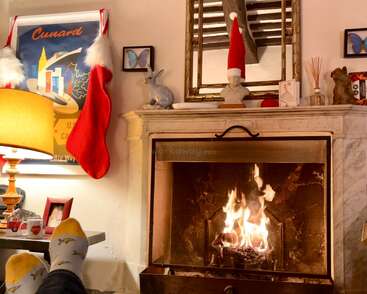 Gemütliches Wohnzimmer mit prasselndem Kaminfeuer, Weihnachtsstrümpfen und festlicher Dekoration. Eine Person, die sich in Pinguin-Socken entspannt und die festliche Atmosphäre sowie das warme Kaminfeuer genießt.