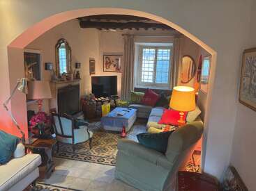 Gemütliches Wohnzimmer mit bunten Kissen, einem Ecksofa, Sesseln, einem Kamin, einem Fernseher und Deko-Kunstwerken. Ein großes Fenster, ein gemusterter Teppich und eine warme Beleuchtung sorgen für eine einladende Atmosphäre.