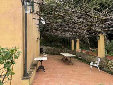 Eine rustikale Terrasse mit Terrakottafliesen, gelben Wänden und einer Pergola, die mit kahlen Ästen bewachsen ist. Tische, Stühle und Pflanzkübel säumen diesen ruhigen, schattigen Bereich.
