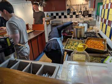 Trois personnes travaillent ensemble dans une cuisine animée. L'une remue le contenu d'une grande marmite, une autre prépare les plats, tandis que les légumes rôtissent sur des plaques, créant une ambiance animée et conviviale.
