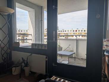 Cette photo montre la vue depuis l'intérieur d'une pièce, à travers des portes vitrées, donnant sur un balcon. On y voit des plantes en pot, un radiateur, une table et des immeubles d'habitation à l'extérieur.