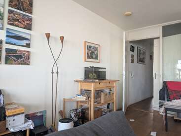 Un salon chaleureux orné de tableaux, avec une étagère en bois sur laquelle trône un aquarium, un grand lampadaire, des livres, des paniers, ainsi qu'un couloir menant aux autres pièces.