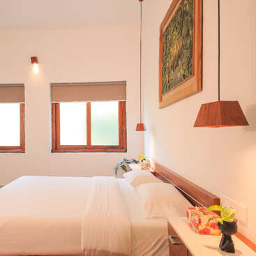 Un dormitorio luminoso y minimalista con paredes blancas, grandes ventanas, una cama perfectamente hecha, detalles en madera, lámparas colgantes, plantas y un cuadro con motivos naturales, que transmite tranquilidad.