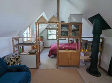 Cette chambre mansardée confortable comprend un lit avec une couverture rouge, des étagères en bois, des guirlandes électriques, des livres, une chaise bleue, un poêle à bois et des plafonds inclinés.