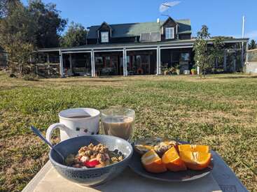Un délicieux petit-déjeuner en plein air composé de granola, de fruits, de tranches d'orange, de café et de thé est servi sur un plateau devant une charmante maison de campagne.