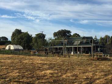 L'image représente une scène rurale sereine, avec une grande maison au toit vert, une balançoire et une serre, sur fond d'arbres et de ciel bleu.