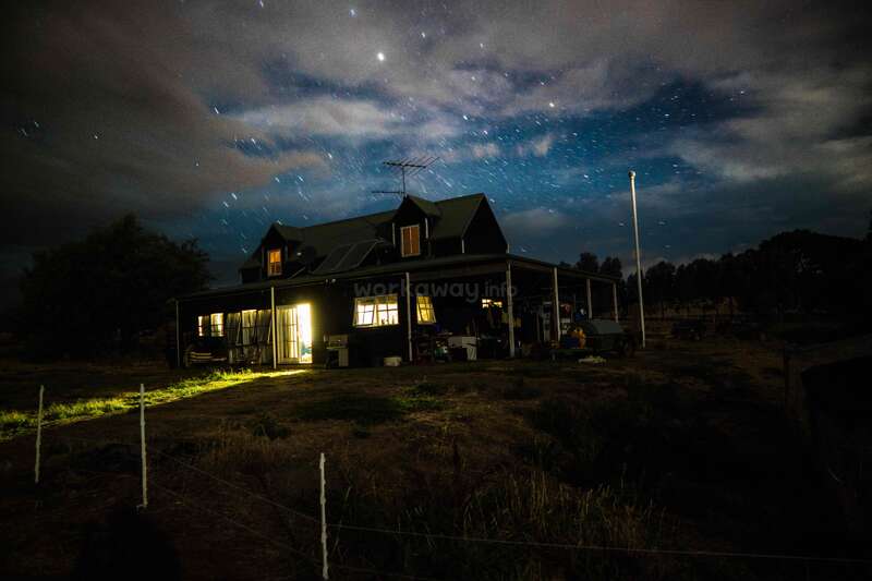 L'image représente une grande maison avec un porche et une cour la nuit, avec un ciel étoilé et une atmosphère nuageuse, le mot "WORK" étant visible sur le côté de la maison.
