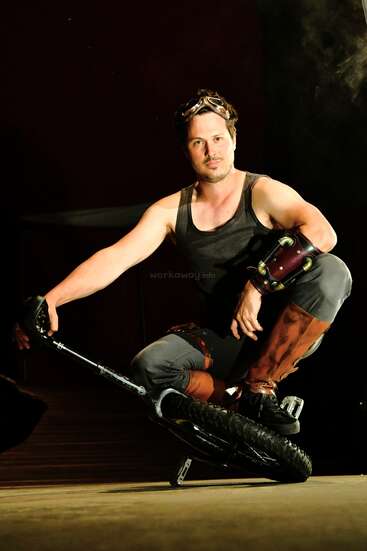 A man poses confidently on a unicycle, wearing a black tank top, brown boots, arm guards, and goggles atop his head, with dramatic lighting behind.