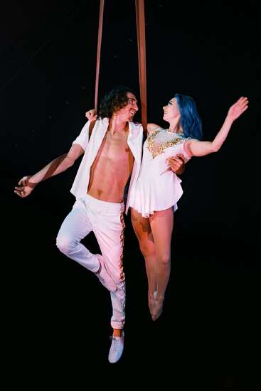 A male and female acrobat perform mid-air on straps. They smile at each other, dressed in white costumes, illuminated against a dramatic, dark background.