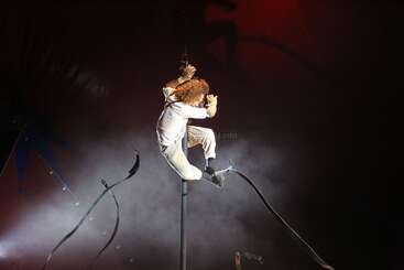 A circus performer with curly hair climbs and balances on a vertical pole, surrounded by theatrical smoke and dramatic lighting, creating a sense of suspense and excitement.