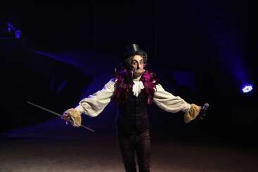 A performer dressed in vintage circus attire, with a feathered collar, top hat, and holding a stick and microphone, stands under dramatic stage lighting.