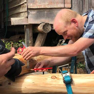 Un hombre está trabajando en una viga de madera, utilizando una herramienta roja para cortarla, con un nivel amarillo y una correa azul cerca, en un entorno rústico al aire libre.
