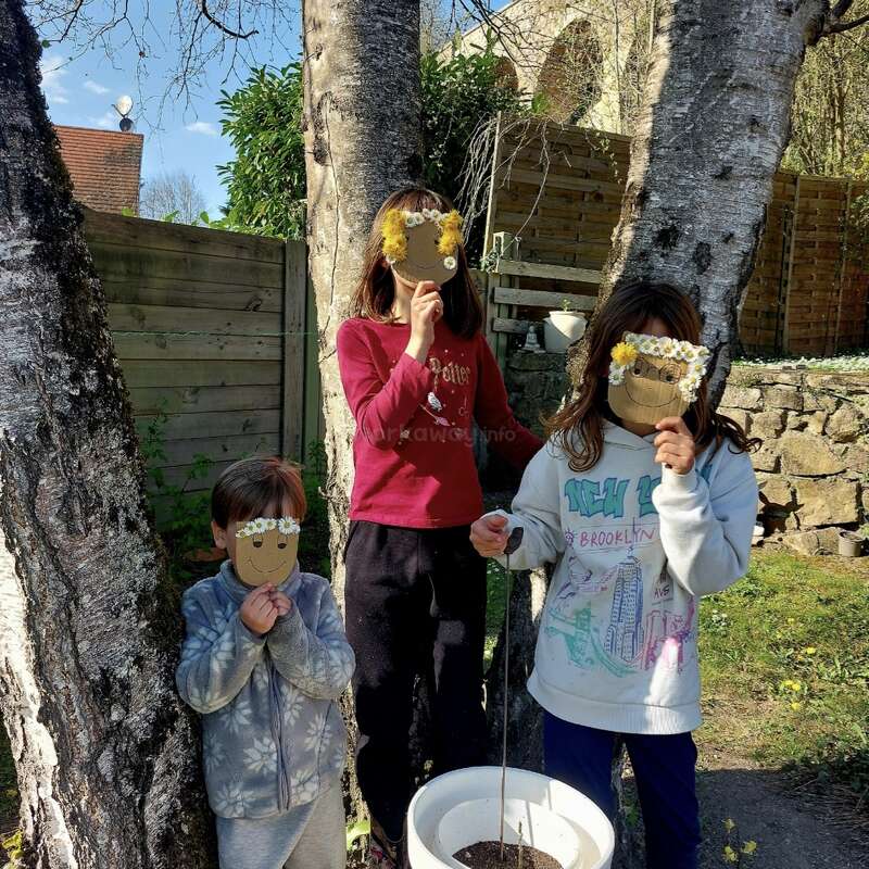 Three children stand outdoors between trees, holding cardboard smiley face masks decorated with flowers. Sunlight filters through, casting shadows. There's a white bucket with soil in front.