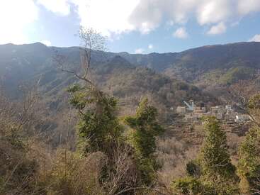 Un petit village est niché à flanc de montagne, entouré d'arbres verts denses et d'un terrain accidenté, sous un ciel partiellement nuageux traversé par la lumière du soleil.