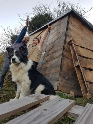 Ein schwarz-weißer Hund sitzt im Vordergrund, während zwei lächelnde Menschen neben einem rustikalen Holzschuppen feiern, mit blattlosen Bäumen und Holzplanken in der Nähe.