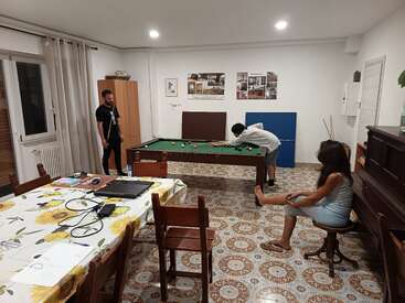 Three people are in a cozy room: two playing pool, one sitting nearby. A table with laptops, a piano, wall art, and patterned floor complete the scene.