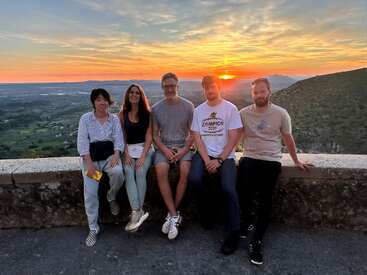 Cinco personas sentadas en un muro de piedra sonríen al atardecer. Detrás de ellos se extiende un pintoresco paisaje con campos, montañas y un colorido cielo nocturno.