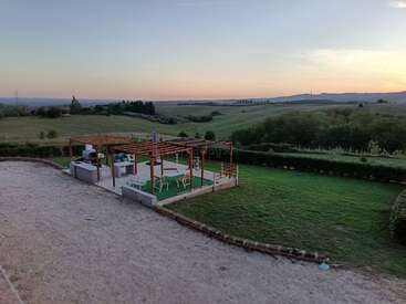 Un apacible paisaje al atardecer con un patio con pérgolas, asientos y zona de barbacoa, con vistas a ondulantes colinas verdes, árboles y campo abierto. Ambiente sereno.