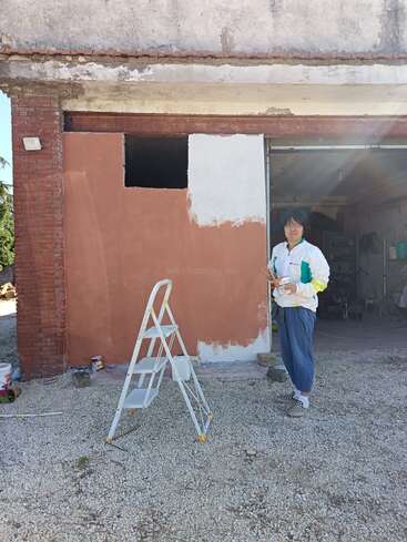 Una persona sujeta un rodillo de pintura junto a una escalera y una pared parcialmente pintada. El edificio parece desgastado. La luz del sol brilla a través de la puerta del garaje abierta detrás de ellos.