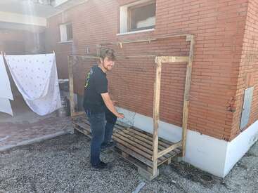 Un hombre sonríe a la cámara junto a una estructura de madera adosada a una pared de ladrillo. Cerca, ropa blanca cuelga de un tendedero.