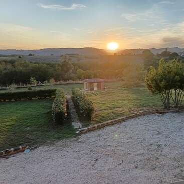 Uma bela cena campestre ao pôr do sol, com um gramado verde, cercas vivas bem aparadas, um pequeno galpão, árvores, área de cascalho e colinas ao fundo.